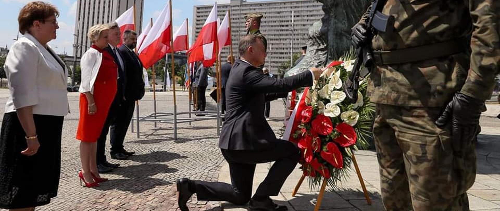Prezydent RP składa wieniec ku pamięci Powstańców Śląskich. Uroczystość w Katowicach. Na zdjęciu klęczący przed pomnikiem prezydent RP, poprawiający szarfę na wieńcu. Wieniec z biało-czerwonych kwiatów. Za prezydentem na baczność w rzędzie stoją cztery osoby. Od lewej starsza kobieta, wicewojewoda opolski oraz dwóch mężczyzn. W tle biało-czerwone flagi. Po prawej stronie kadru stoi żołnierz, który pełni wartę przy pomniku.