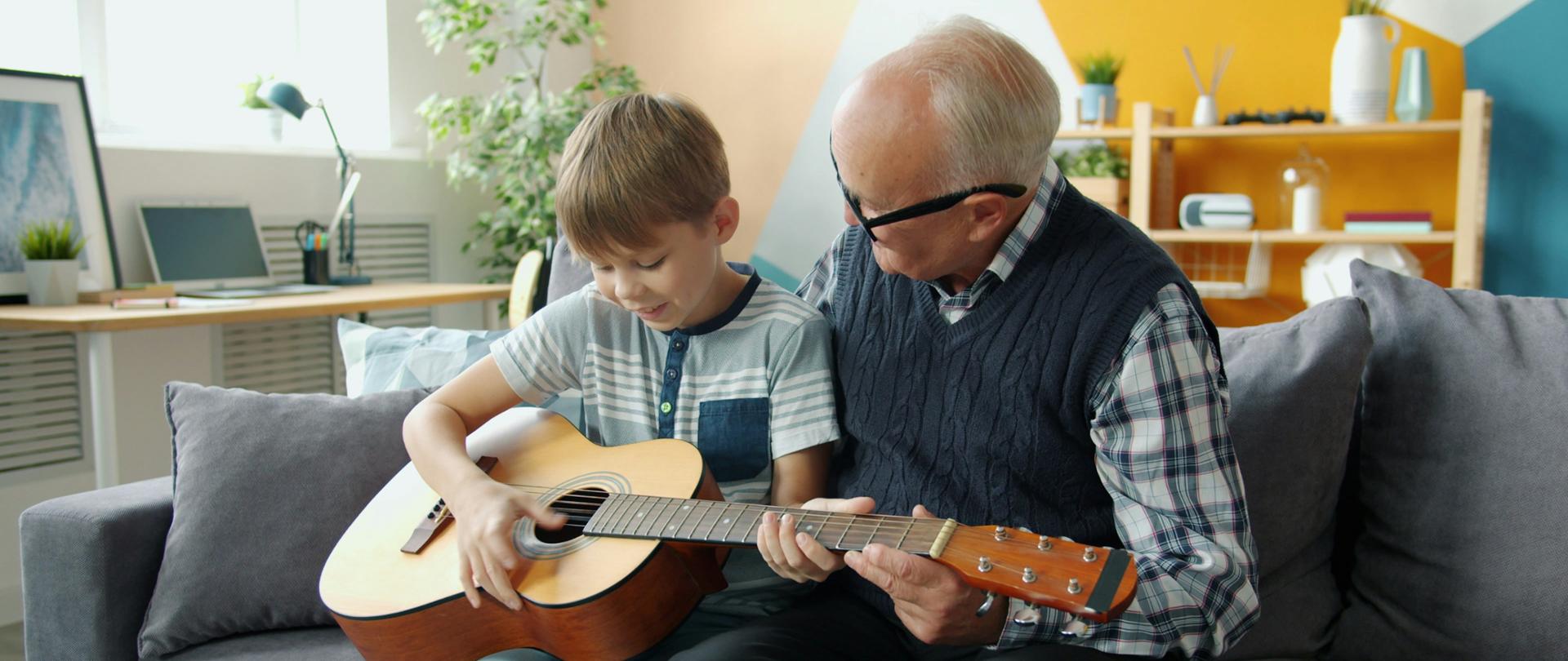Wnuk uczy się grać na gitarze akustycznej z pomocą dziadka, siedząc na kanapie w domu.