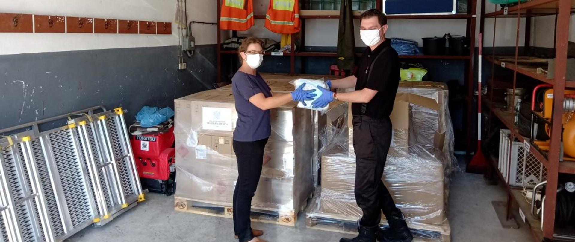 Zdjęcie zrobione w pomieszczeniu magazynowym Komendy Powiatowej Państwowej Straży Pożarnej w Lesku. Na zdjęciu na pierwszym planie stoi przedstawicielka jednej ze szkół powiatu leskiego odbierająca z rąk funkcjonariusza Komendy Powiatowej Państwowej Straży Pożarnej pakiety maseczek ochronnych. W tle dwójki osób widać palety ze środkami ochrony indywidualnej dla pozostałych szkół z terenu powiatu leskiego.