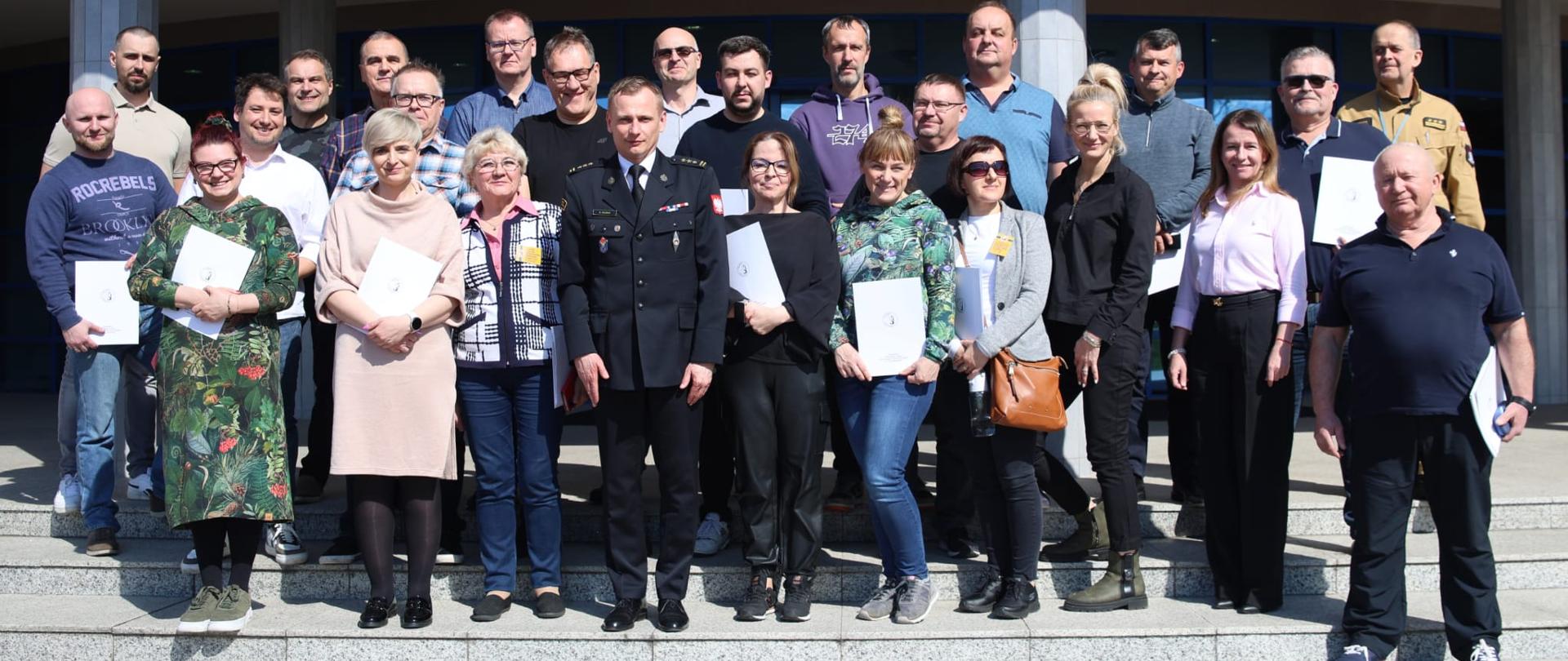 Pamiątkowe zdjęcie grupowe uczestników szkolenia aktualizującego dla inspektorów ochrony przeciwpożarowej (23 osoby) w towarzystwie Zastępcy Komendanta Centralnej Szkoły PSP, oficera CS PSP oraz kobiety