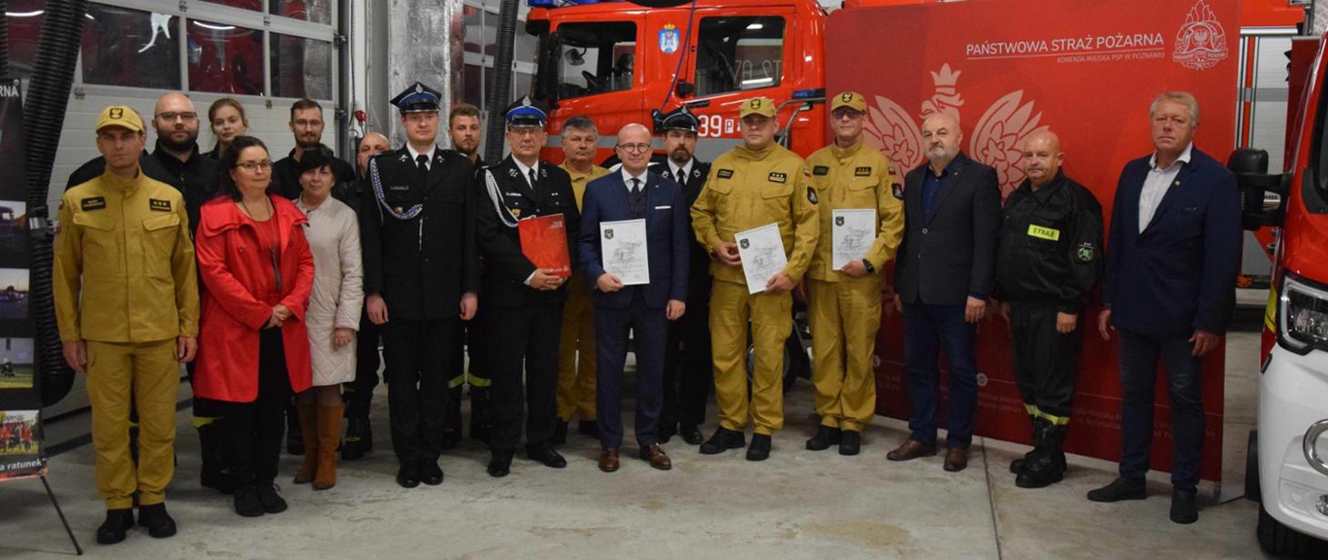 Zdjęcie przedstawia strażaków PSP i OSP oraz zaproszonych gości, którzy uczestniczyli w przekazaniu łodzi dla OSP Głuszyna. W tle wóz strażacki i ścianka PSP.