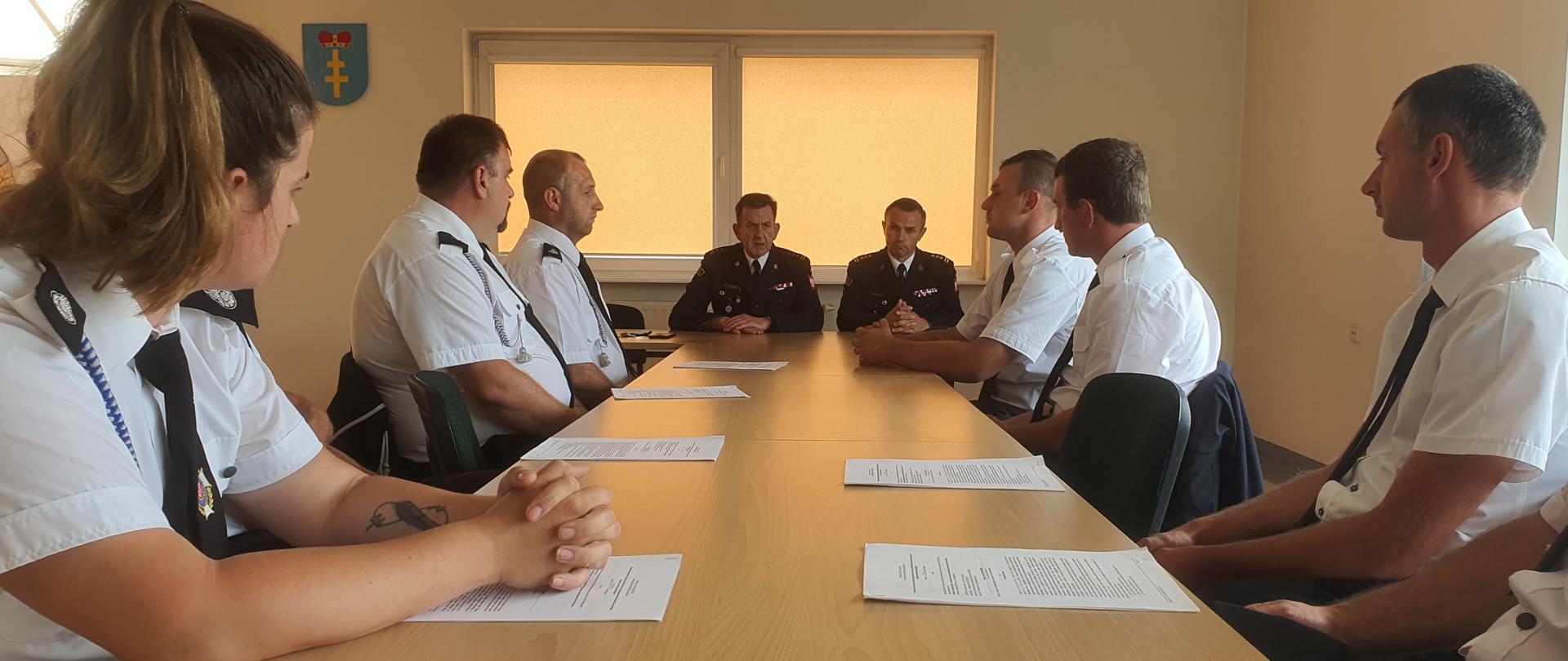 Na zdjęciu widzimy przy stole prezydialnym dwóch mężczyzn w mundurach PSP. Rozmawiają z siedzącymi przed nimi strażakami OSP. Przed strażakami OSP leża dokumenty.