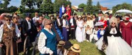 grupa uczestników wydarzenia, w strojach barokowych, pozują do zdjęcia na otwartej przestrzeni 