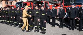 Strażak w mundurze koloru musztardowego wita się ze strażakami z jednostki ratowniczo-gaśniczej za nimi stoją strażacy ochotniczych straży pożarnych oraz ustawione są samochody pożarnicze.