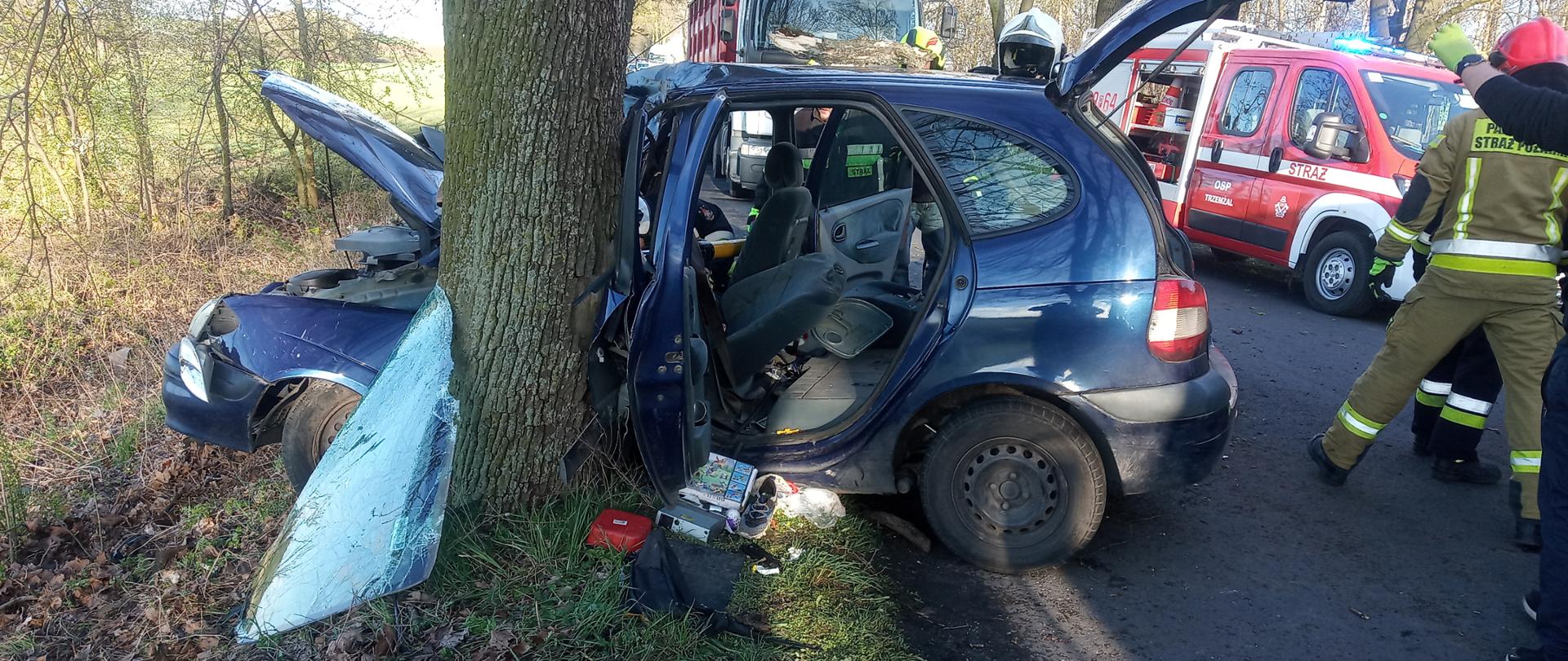 Na zdjęciu widoczny jest samochód osobowy, który uderzył w przydrożne drzewo. Przylega on bezpośrednio do drzewa na wysokości drzwi kierowcy. Widoczne uszkodzenia pojazdu, wybita przednia szyba. Po prawej stronie widać strażaków a w tle czerwony samochód strażacki