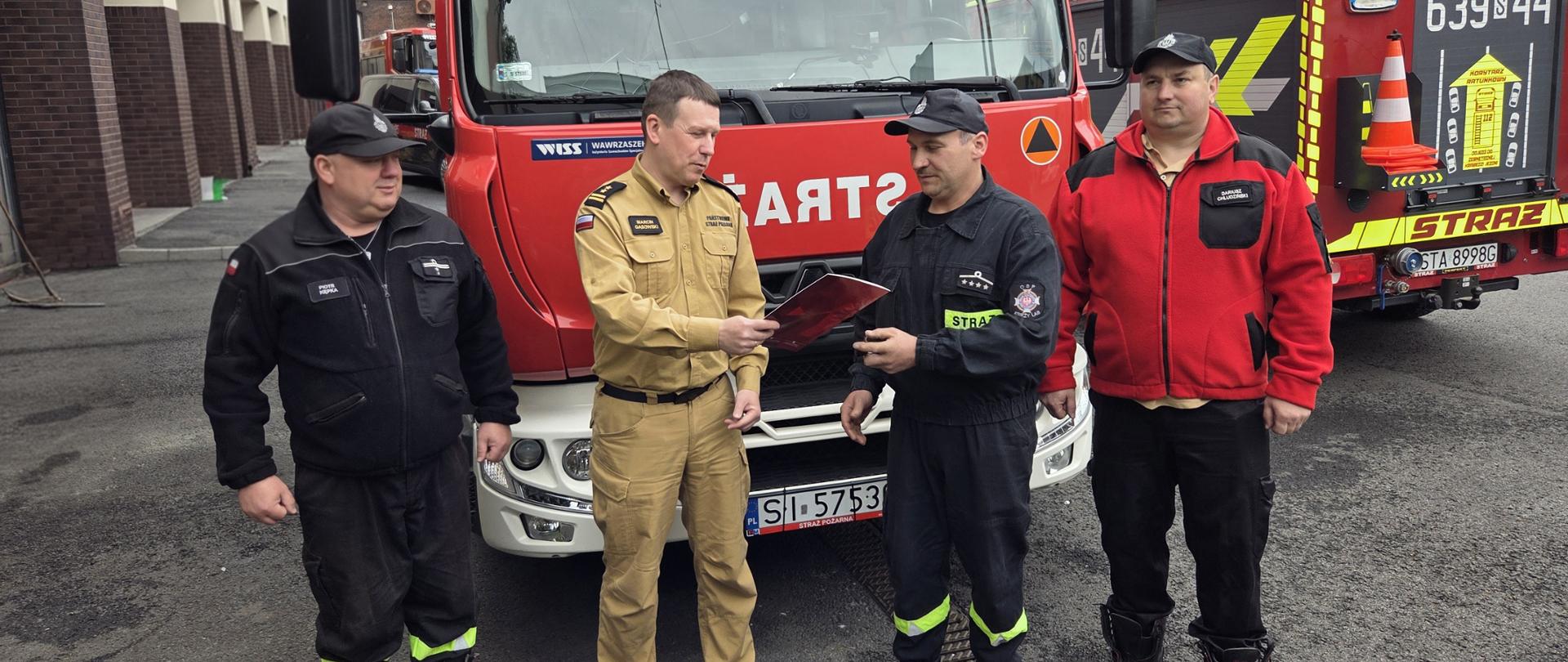 Na zdjęciu widać czterech strażaków stojących przed czerwonym samochodem ratowniczo-gaśniczym PSP. W centrum fotografii znajduje się komendant miejski PSP w Siemianowicach Śląskich, bryg. Marcin Gąsowski, ubrany w jasny, służbowy mundur. Wręcza on czerwoną teczkę prezesowi OSP Księży Las Adamowi Skowronowi, który stoi obok w czarnym ubraniu strażackim.
Pozostali strażacy stoją po bokach, a w tle widoczne są kolejne wozy strażackie oraz zabudowania jednostki straży pożarnej. Zdjęcie przedstawia oficjalny moment przekazania dokumentów przekazania samochodu ratowniczo-gaśniczego dla OSP Księży Las. Wydarzenie odbyło się w strażackiej atmosferze służbowej.