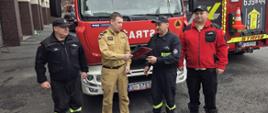 Pozostali strażacy stoją po bokach, a w tle widoczne są kolejne wozy strażackie oraz zabudowania jednostki straży pożarnej. Zdjęcie przedstawia oficjalny moment przekazania dokumentów przekazania samochodu ratowniczo-gaśniczego dla OSP Księży Las. Wydarzenie odbyło się w strażackiej atmosferze służbowej.