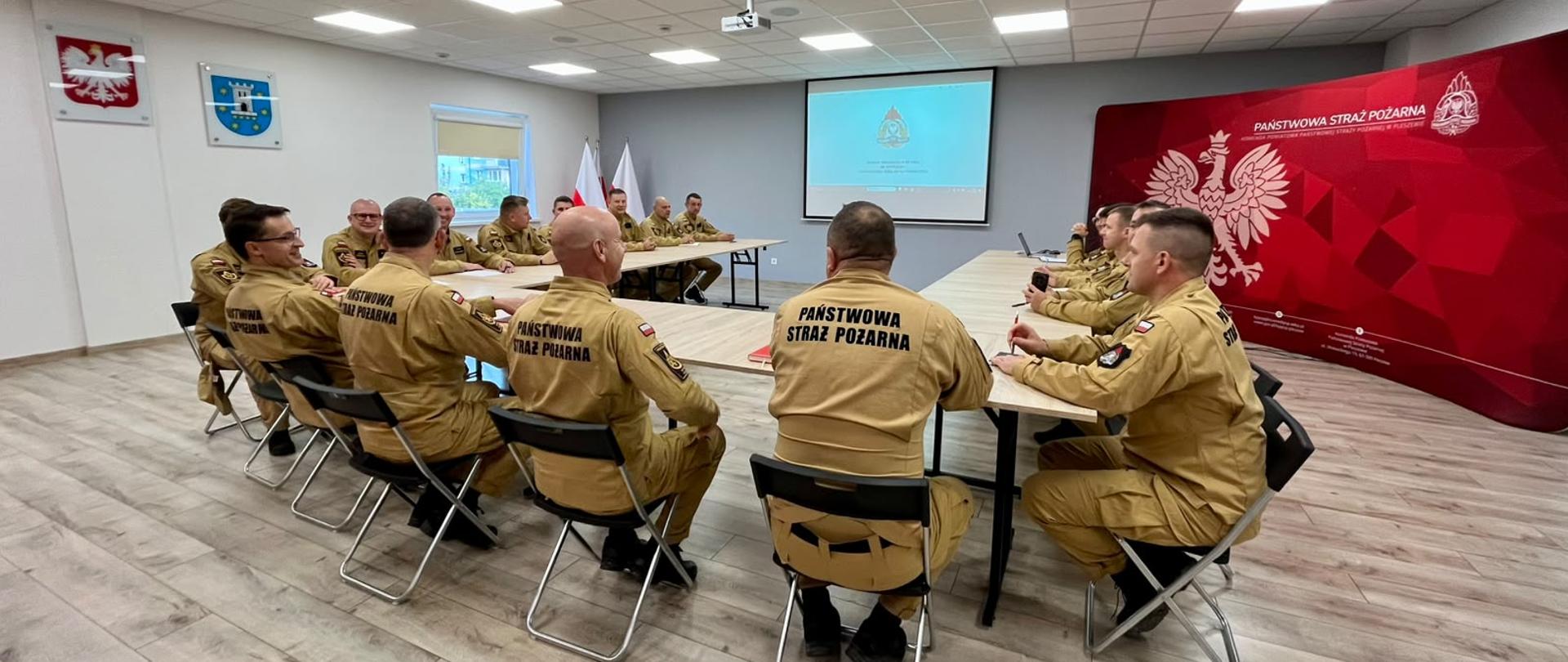 Strażacy w piaskowych mundurach służbowych Państwowej Straży Pożarnej siedzą przy stołach ustawionych w podkowę podczas odprawy w sali konferencyjnej. Na ścianie po prawej stronie widoczne jest czerwone tło z godłem Polski i napisem „Państwowa Straż Pożarna”, a w tle znajduje się ekran projekcyjny oraz polskie flagi.