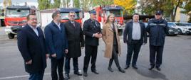 na zdjęciu sześciu mężczyzn i jednak kobieta, kobieta ubrana w jasny płaszcz, jeden z mężczyzn to komendant wojewódzki PSP w łodzi w mundurze wyjściowym pozostali ubrani w płaszcze i kurtki, w tle samochody straży pożarnej i drzewa