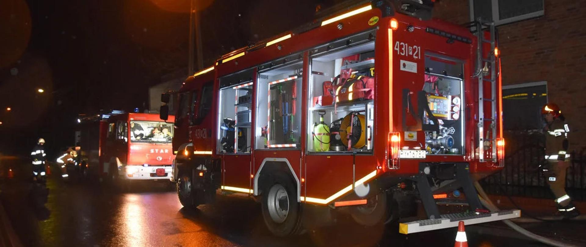 Zdjęcie przedstawia dwa wozy strażackie. Wozy stoją na drodze publicznej przed budynkami domów jednorodzinnych. Samochody są oświetlone. W jednym samochodzie wszystkie skrytki na sprzęt pożarniczy są otwarte, podawana jest z niego woda potrzebna do ugaszenia pożaru.