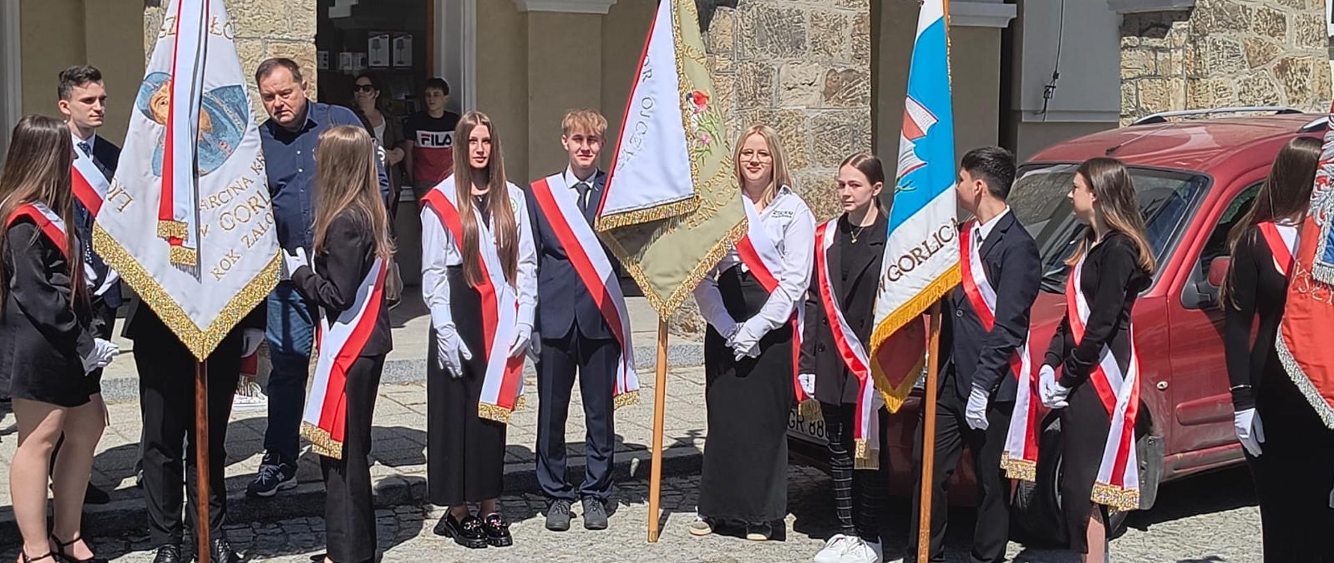 Grupa uczniów z pocztów sztandarowych ustawiona przed arkadami kamienicy na rynku.