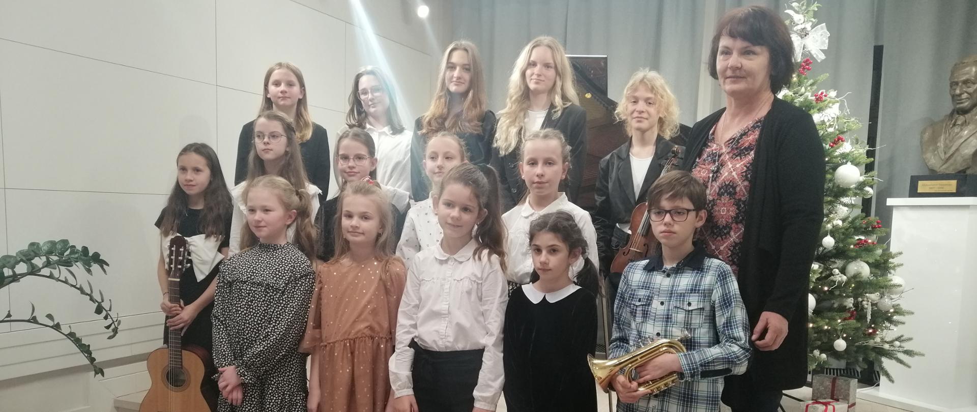 Zdjęcie przedstawia uczniów stojących w trzech rzędach na stopniach sceny. Z prawej strony stoi nauczyciel, a przed nim uczeń trzymający trąbkę. Za nauczycielem widać ubraną choinkę z prezentami a tuż obok niej postument z popiersiem patrona szkoły. Od lewej strony stoi dziewczynka trzymająca gitarę. W tle widać zasunięte kotary w szarym kolorze. 