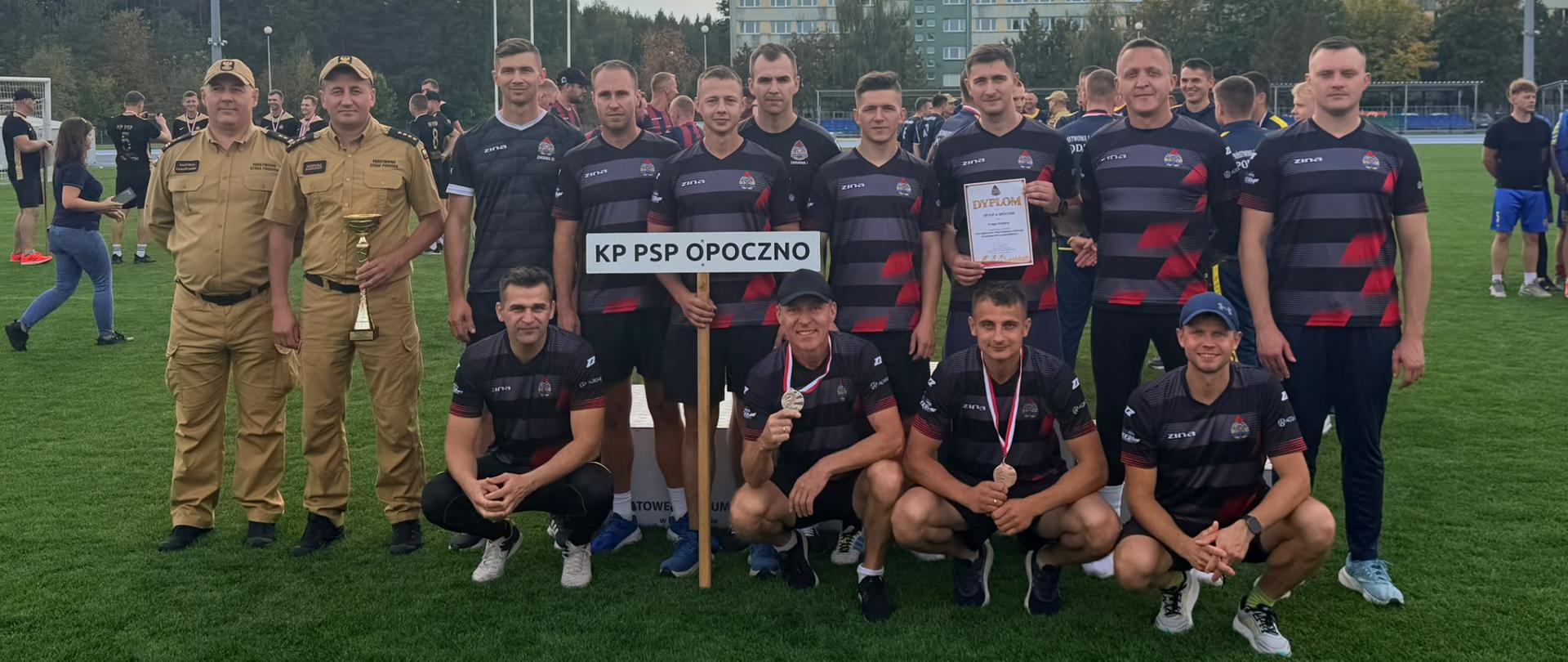 2. Na zdjęciu drużyna Komendy Powiatowej Państwowej Straży Pożarnej w Opocznie pozuje do wspólnej fotografii na stadionie Powiatowego Centrum Sportu w Bełchatowie. Na pierwszym planie stoi grupa strażaków ubranych w jednakowe czarne koszulki sportowe z czerwonymi i szarymi pasami oraz czarne spodenki. Większość zawodników stoi, natomiast czterech mężczyzn kuca z przodu. Dwaj zawodnicy mają na szyi medale, jeden trzyma dyplom, a kolejny trzyma tabliczkę z napisem „KP PSP OPOCZNO”. Po lewej stronie grupy stoją dwaj strażacy w piaskowych mundurach służbowych. Jeden z nich trzyma puchar. W tle widać innych uczestników zawodów, zieloną murawę stadionu, drzewa oraz budynek. Nad obiektem rozciąga się zachmurzone niebo z prześwitami światła słonecznego.