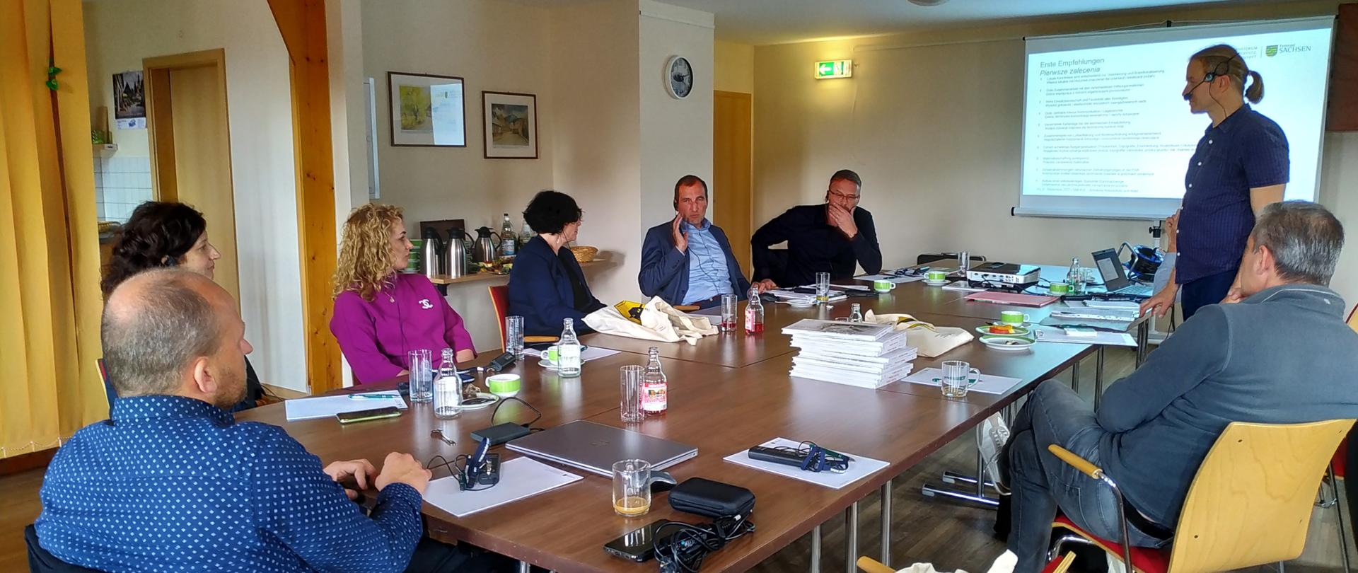 W sali za stołem siedzi grupa kilku kobiet i mężczyzn. Po prawej stronie na ścianie wisi ekran z wyświetloną prezentacją.