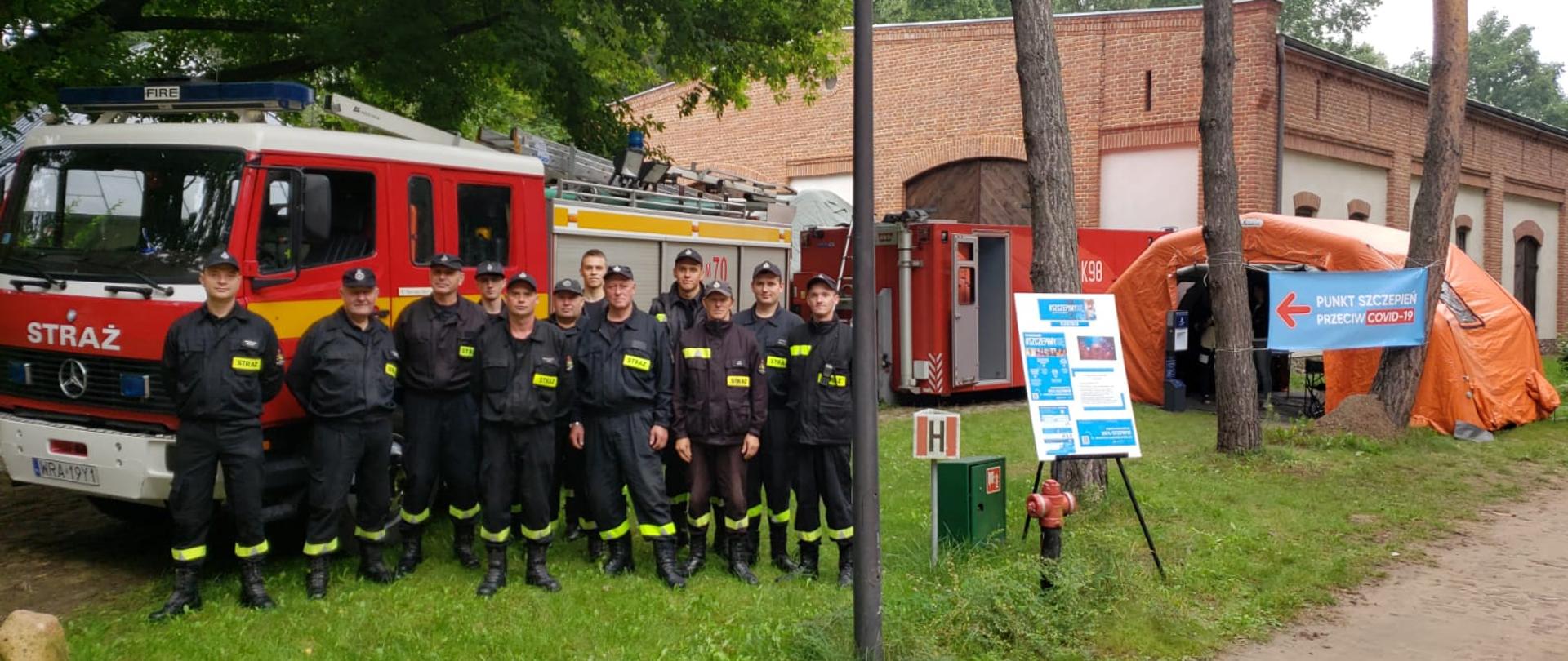 Zdjęcie przedstawia plenerowy punkt szczepień utworzony na terenie Muzeum Wsi Radomskiej podczas Dożynek Województwa Mazowieckiego. Na zdjęciu widać od lewej strażaków z Ochotniczej Straży Pożarnej w Kowali. Strażacy ubrani są w ubranie koszarowe w kolorze czarnym z odblaskowymi elementami. Za nimi ustawiony jest pojazd ratowniczo - gaśniczy w kolorze czerwonym z białymi i srebrnymi elementami zabudowy. Po prawej stronie ustawiony jest pompowany namiot pneumatyczny w kolorze pomarańczowym. Za namiotem, w oddali znajduje się ustawiony specjalny kontener sanitarny. Zabudowa kontenera w kolorze czerwonym ze srebrnymi elementami zabudowy. 
