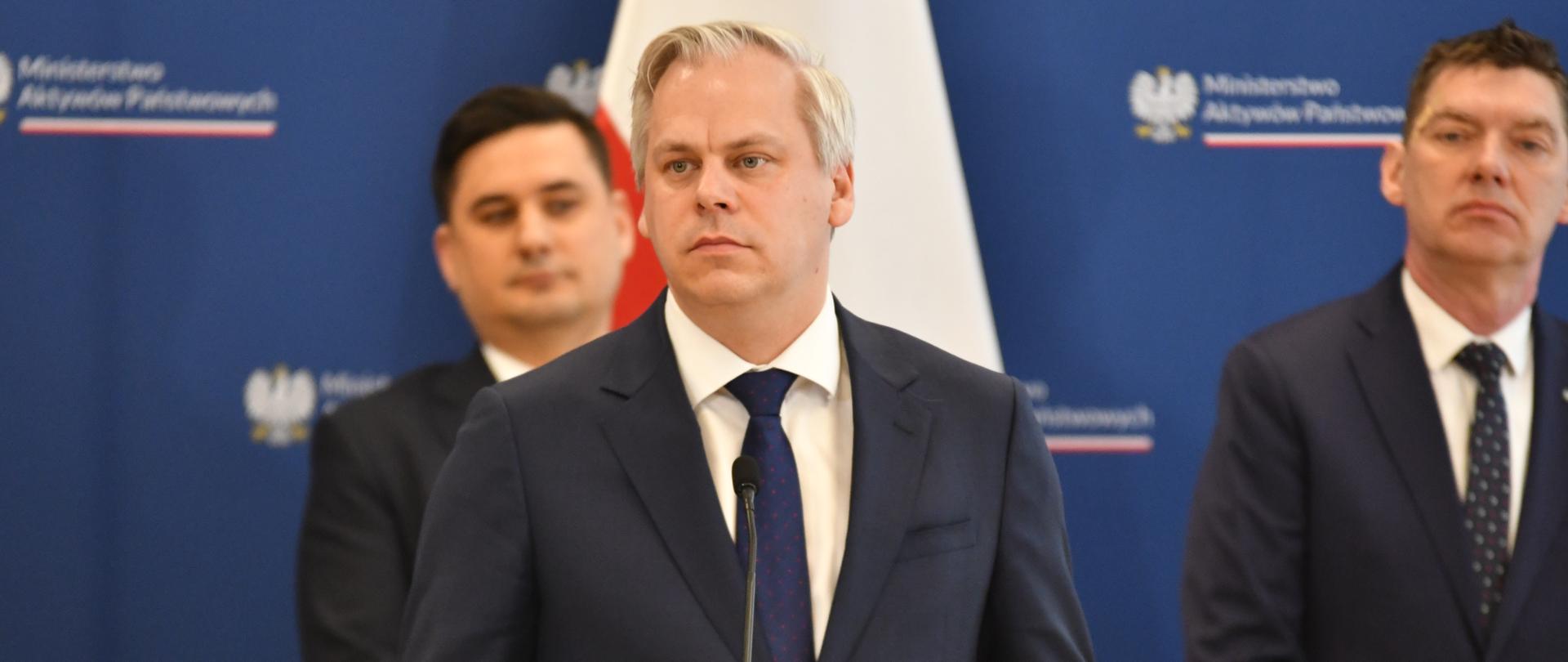 Wiceminister Karol Rabenda stoi za mównicą. W tle pozostali uczestnicy briefingu i ścianka z logotypami MAP
