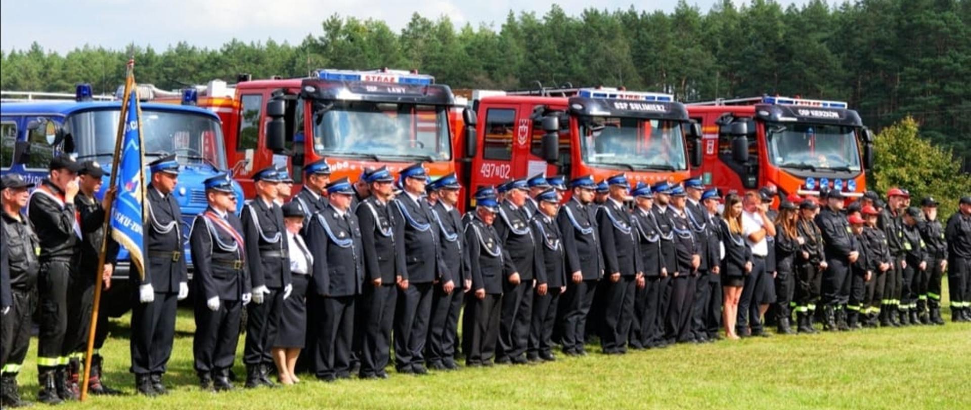 Na zdjęciu Strażacy OSP w umundurowaniu wyjściowym ze sztandarem na tle samochodów pożarniczych