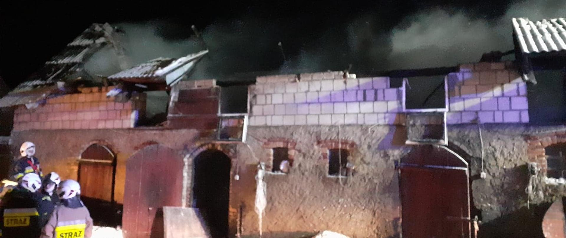 W centrum widać murowany budynek częściowo bez dachu, z którego wydobywają się kłęby dymu. Po lewej stronie widać strażaków.