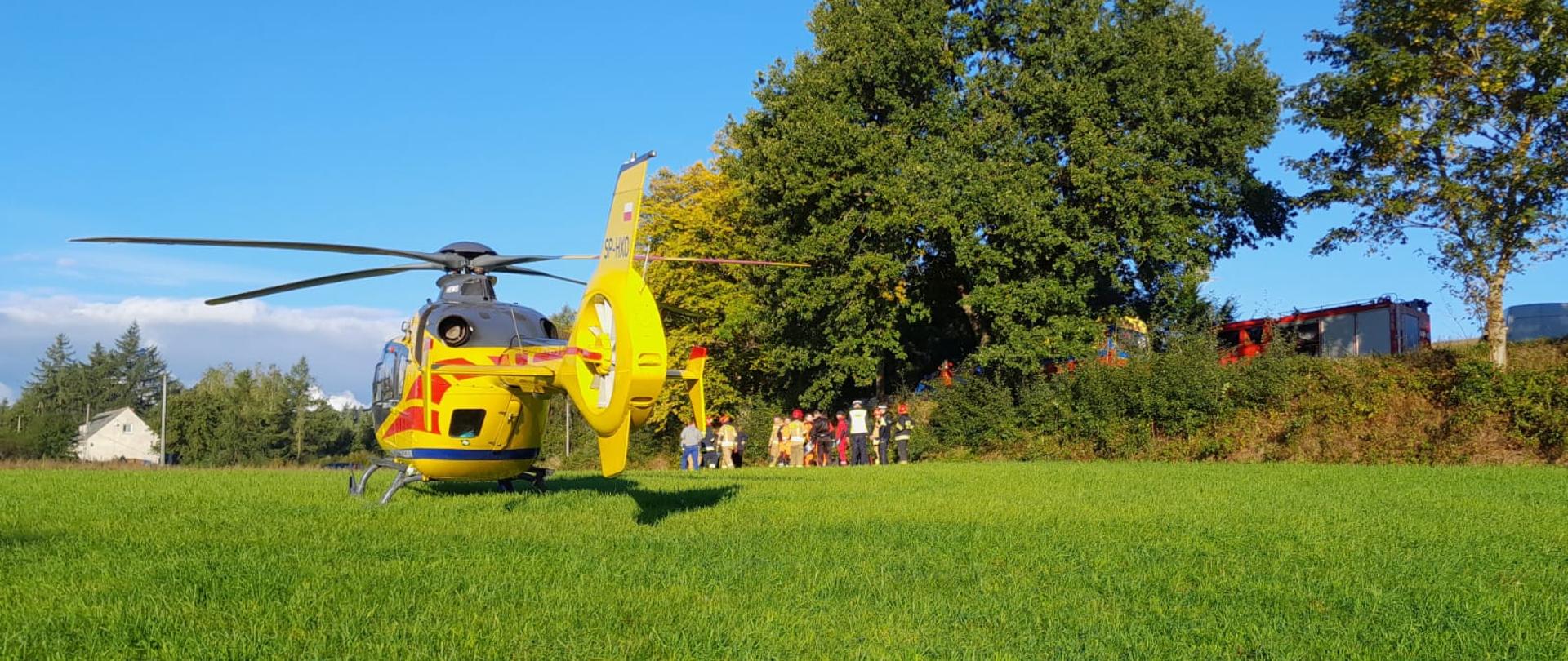 Śmigłowiec LPR, za którym w tle stoją ratownicy przy poszkodowanym.