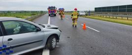 Droga ekspresowa S5. Na jezdni widoczne trzy pojazdy straży pożarnej oraz strażacy rozstawiający pachołki ostrzegawcze. Na pierwszym planie - fragment auta osobowego, stojącego częściowo na pasie awaryjnym. Z kolei na pasie zieleni stoi drugie auto osobowe. Obok niego dwoje ludzi. 