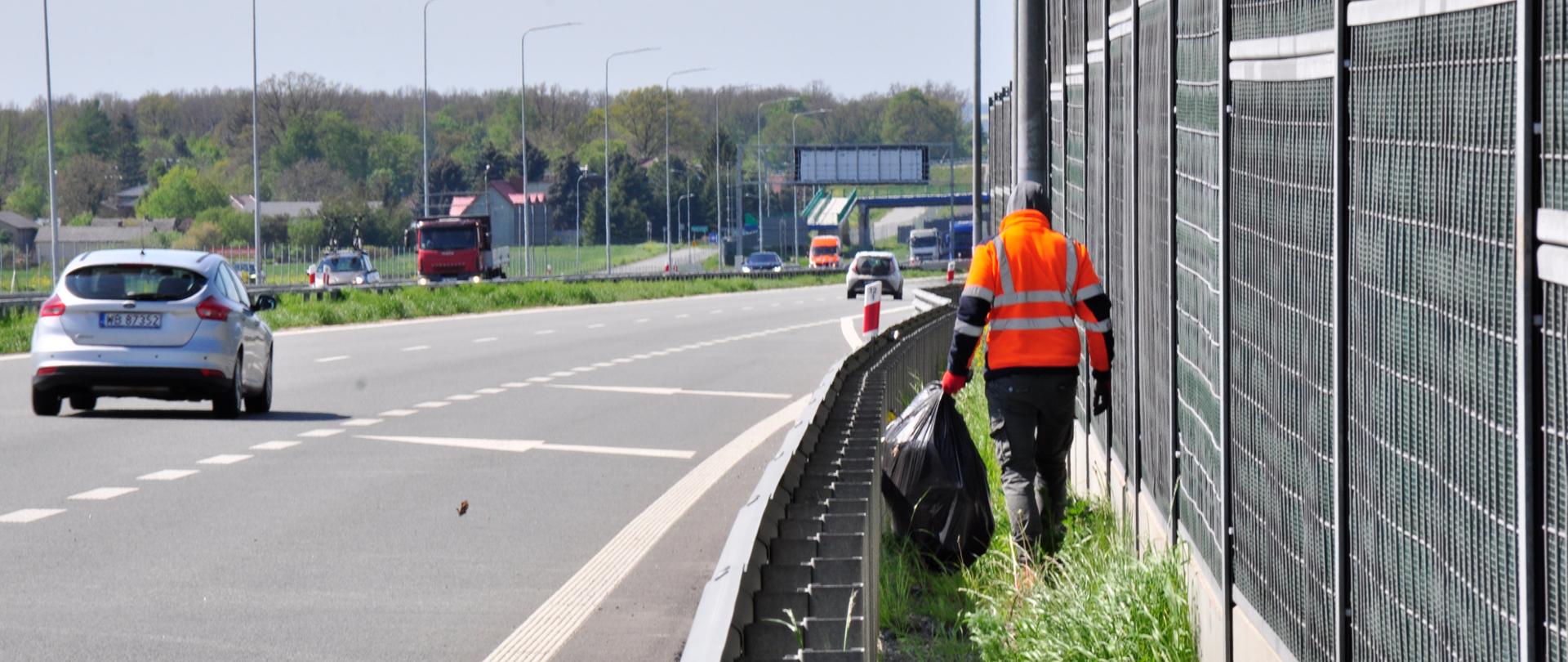 Zdjęcie przedstawia osobę ubraną w pomarańczową kamizelkę odblaskową, która porusza się wzdłuż pobocza autostrady, zbierając śmieci. Osoba ta trzyma duży czarny worek na śmieci i idzie obok metalowego ogrodzenia dźwiękochłonnego. Na drodze w tle widoczne są pojazdy, w tym srebrny samochód osobowy oraz czerwona ciężarówka. Po lewej stronie zdjęcia znajduje się pas zieleni oddzielający jezdnię od pobocza. W tle widoczne są drzewa oraz budynki, a dzień jest pogodny, z lekkim zachmurzeniem.