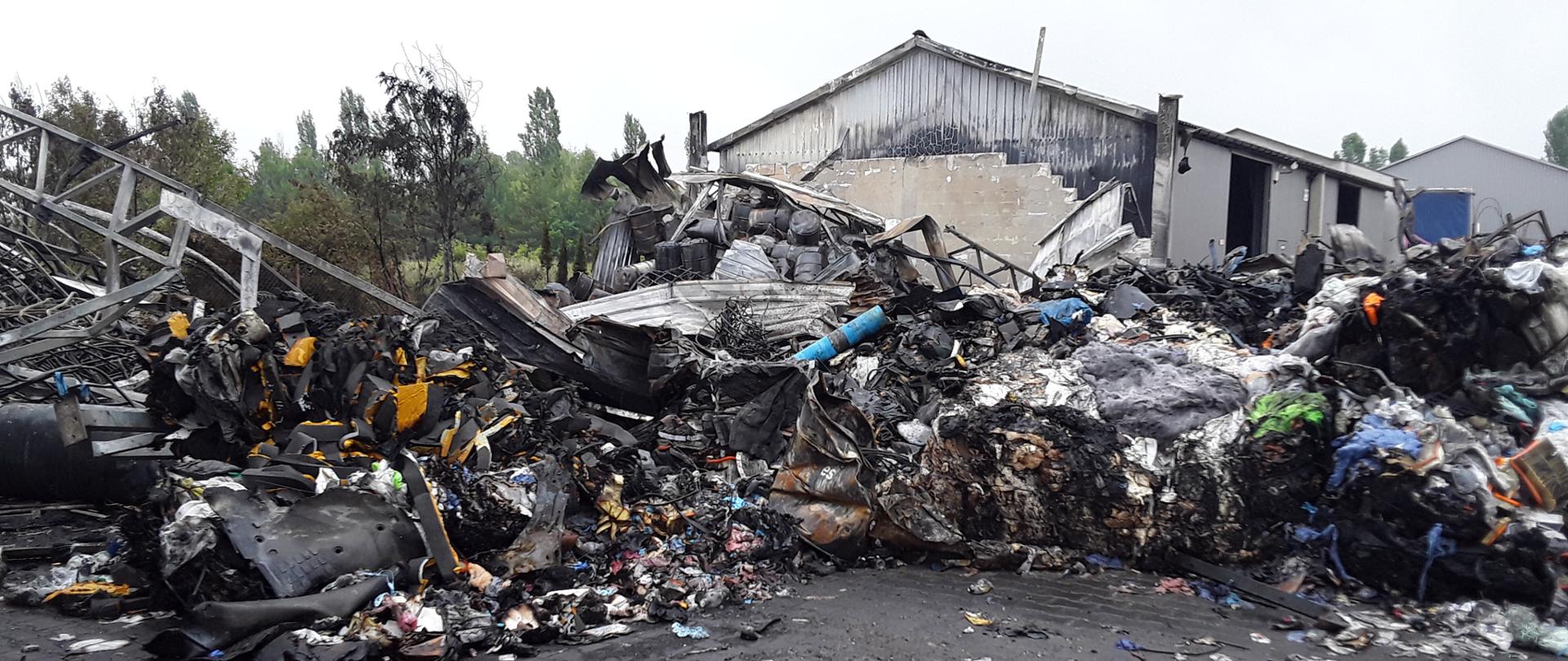 Spalone odpady znajdujące się na otwartym terenie. W tle hala magazynowa.