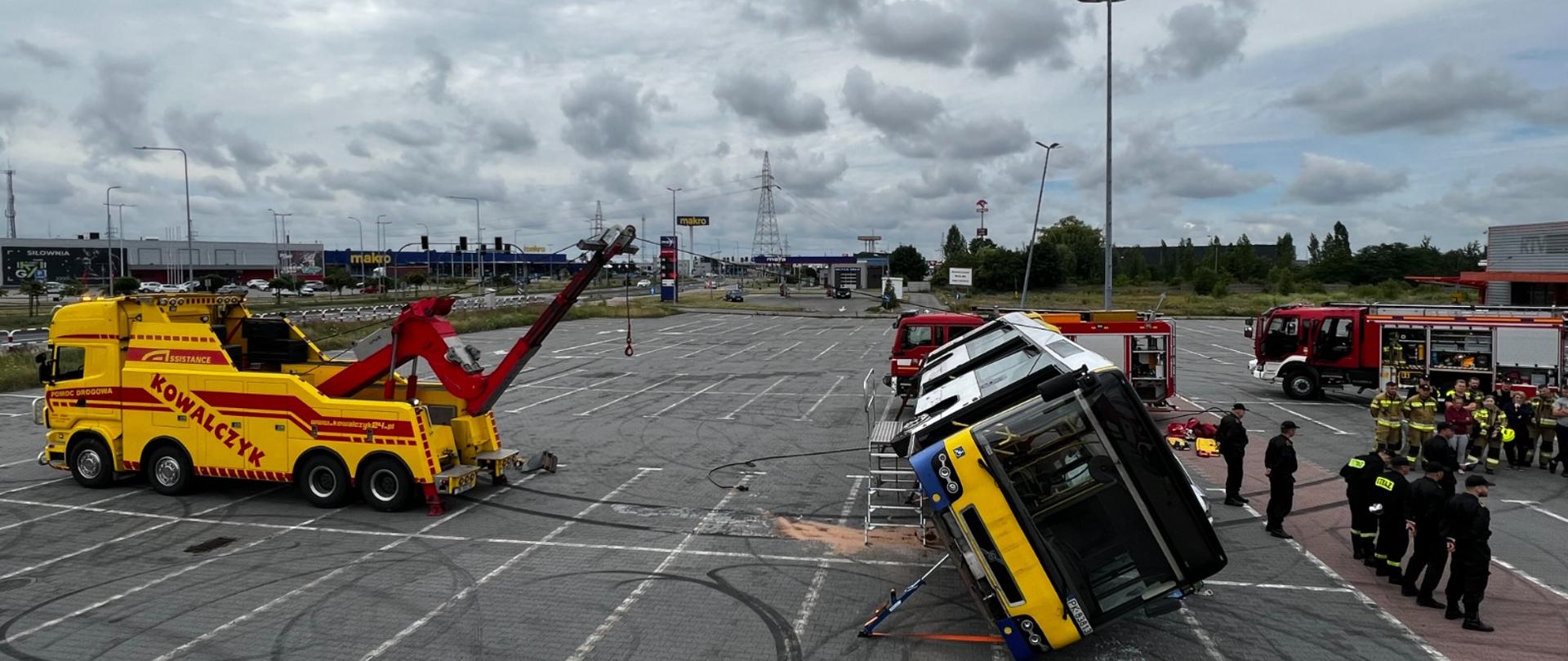 Na zdjęciu widać parking z kostki brukowej. Na środku znajduje się autobus komunikacji miejskiej. Jest pochylony. Po lewej stronie żółty samochód pomocy drogowej. Niebo zachmurzone. W dalszej perspektywie samochody straży pożarnej w kolorze czerwonym. Na parkingu latarnie oświetleniowe.