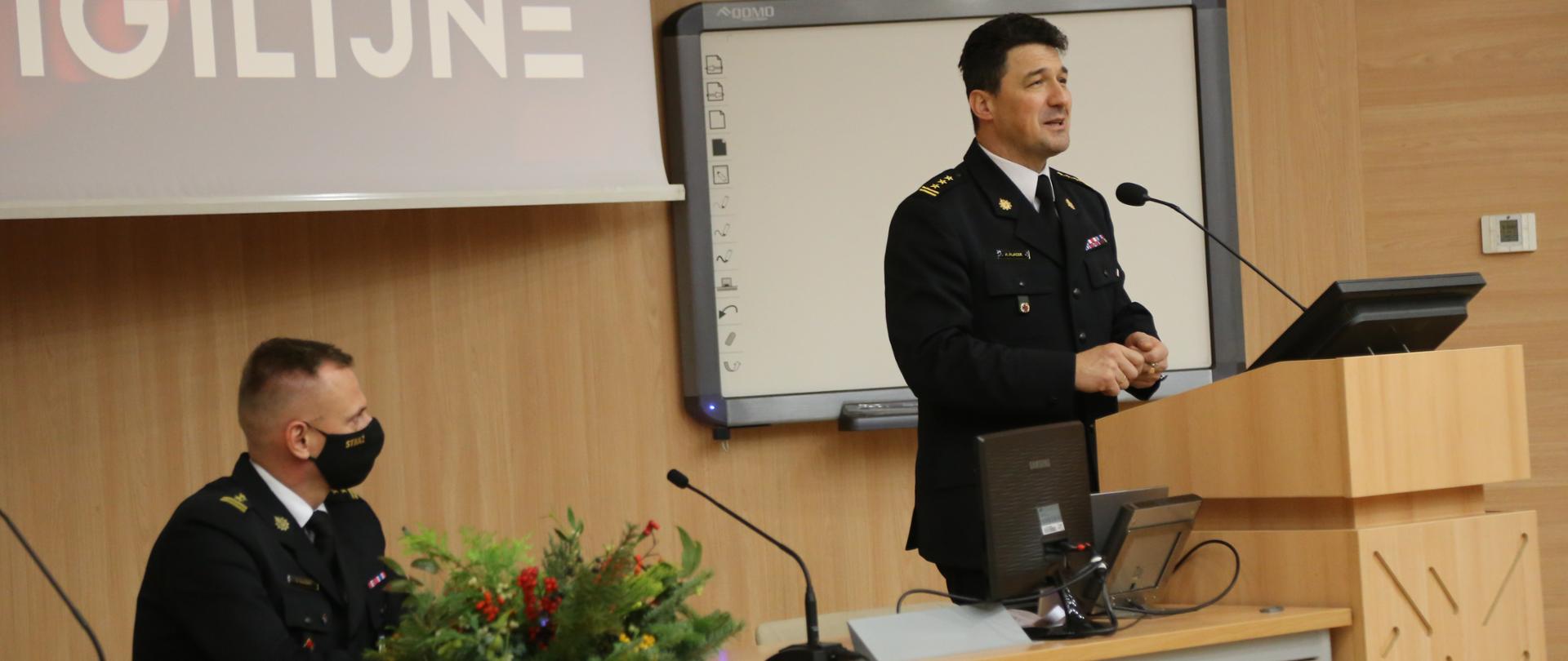 Komendant Centralnej Szkoły PSP przemawia stojąc na mównicy, obok siedzący przy biurku Zastępca Komendanta Centralnej Szkoły PSP. Na biurku świąteczny stroik