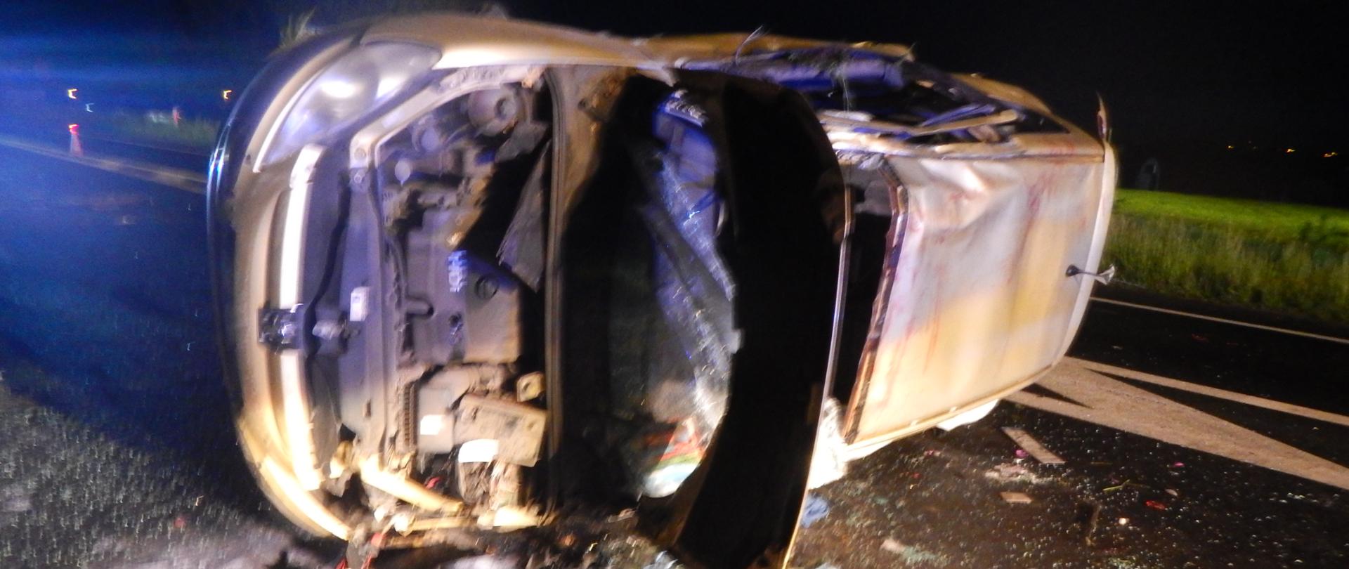 Zdjęcie przedstawia srebrny samochód po wypadku drogowym. Samochód leży na drodze na lewym boku.