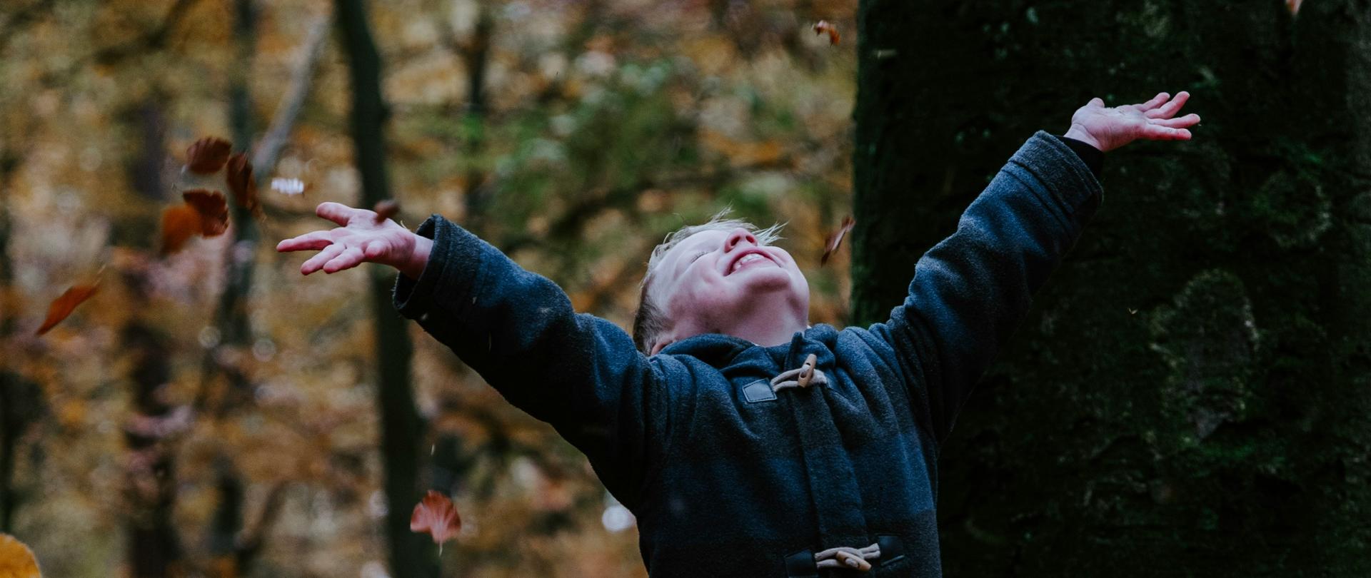 Chłopiec w ciepłym płaszczu stoi w lesie, z radością rozrzucając opadłe liście dookoła siebie. W tle widać drzewo, a obok niego czarnego psa, który węszy wśród liści. Scena jest pełna ciepłych jesiennych barw, a cała atmosfera emanuje beztroską radością.