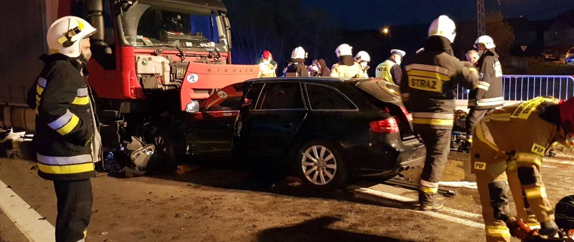 Miejsce zderzenia. Na jezdni widoczne dwa samochody, w tym jeden ciężarowy, które zderzyły się czołowo. Dookoła pojazdów pracują strażacy. 