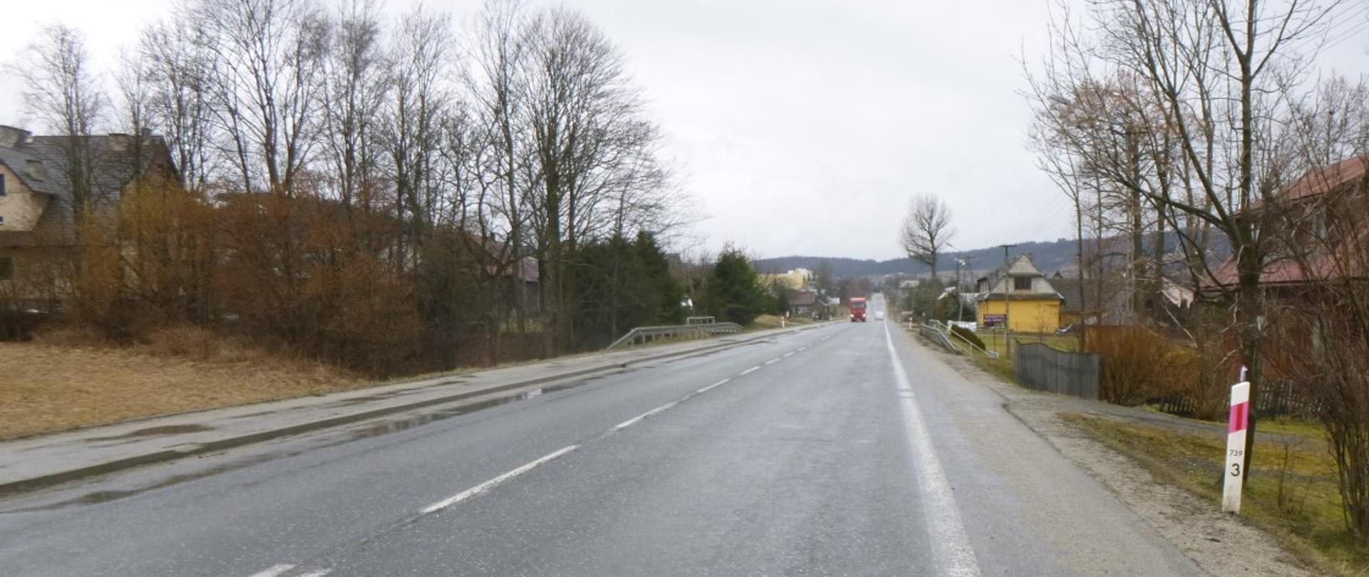 droga prowadząca przez teren zabudowany, blisko drogi roślinność, w oddali samochód ciężarowy