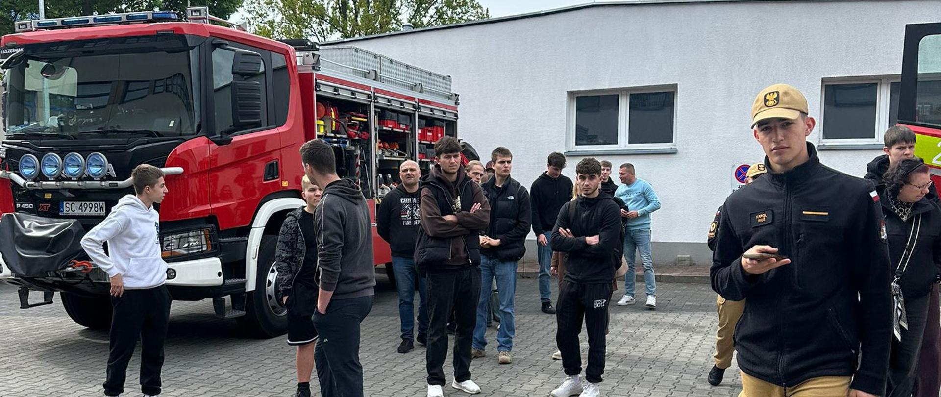 Grupa młodych osób uczestniczących w Dniu Otwartym spaceruje po terenie Centralnej Szkoły PSP pod opieką kadetów w umundurowaniu. Uczestnicy zwiedzania znajdują się przed jednym z budynków na terenie Szkoły oraz oglądają stojący przy nim wóz bojowy CS PSP