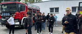 Grupa młodych osób uczestniczących w Dniu Otwartym spaceruje po terenie Centralnej Szkoły PSP pod opieką kadetów w umundurowaniu. Uczestnicy zwiedzania znajdują się przed jednym z budynków na terenie Szkoły oraz oglądają stojący przy nim wóz bojowy CS PSP