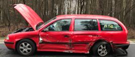 Zdjęcie przedstawia czerwony pojazd marki Skoda z uszkodzonym lewym bokiem i otwartą maską. Samochód stoi na drodze asfaltowej na tle lasu. 