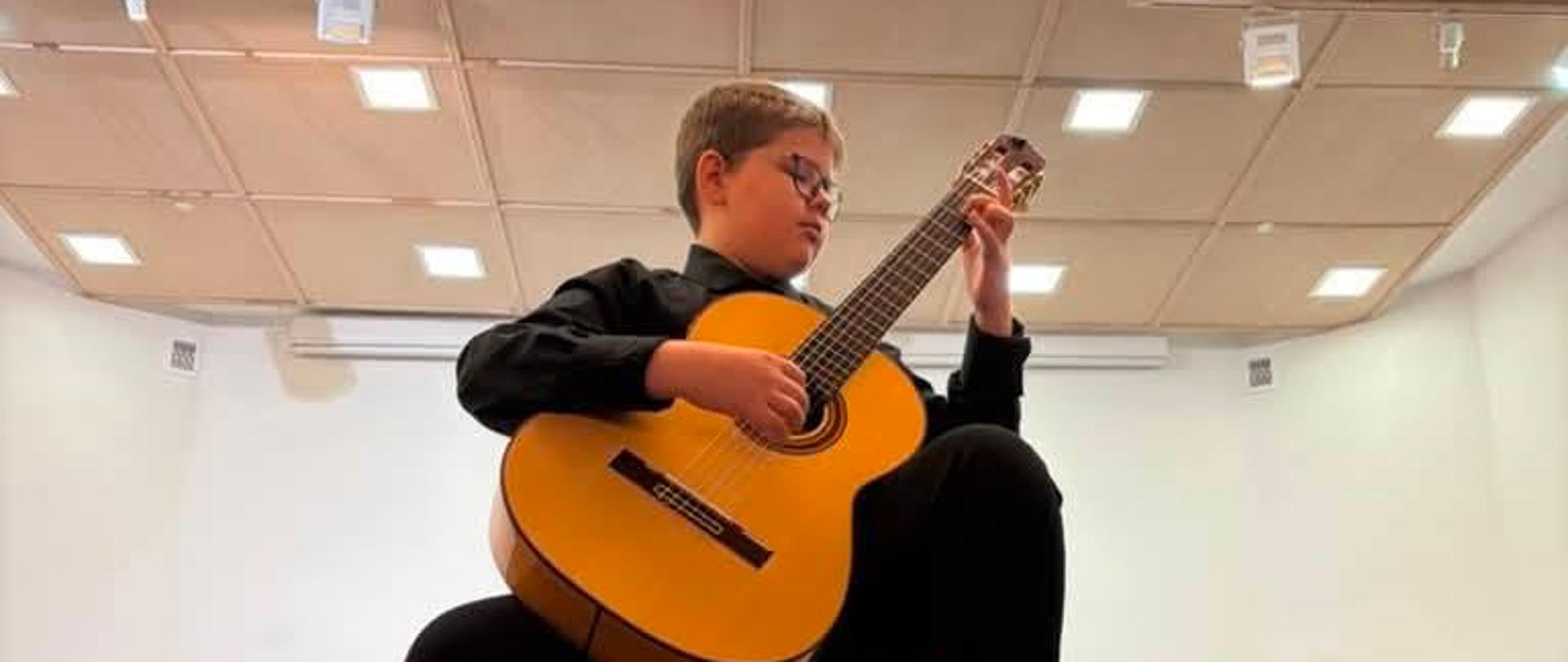 W sali koncertowej na taborecie siedzi chłopiec ubrany w czarne spodnie i czarną koszulę. Chłopiec gra na gitarze klasycznej. Jedną nogę ma ustawioną na podnóżku.
