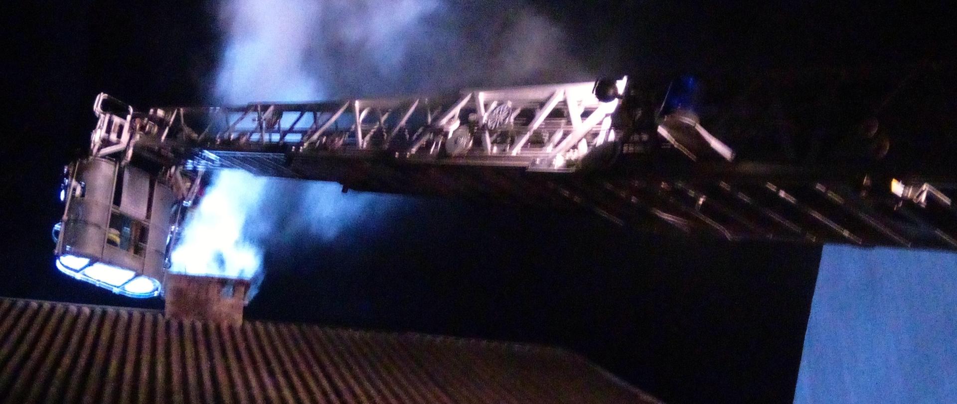 Zdjęcie przedstawia strażaków gaszących pożar sadzy w kominie z kosza drabiny mechanicznej. W tle widać wydobywający się dym z komina. Dach budynku jest oświetlony