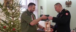 mężczyzna harcerz w zielonym mundurze przekazuje lampion z zapaloną świeczką strażakowi w ciemnym mundurze , w tle choinka z biał oczerwonymi lampkami