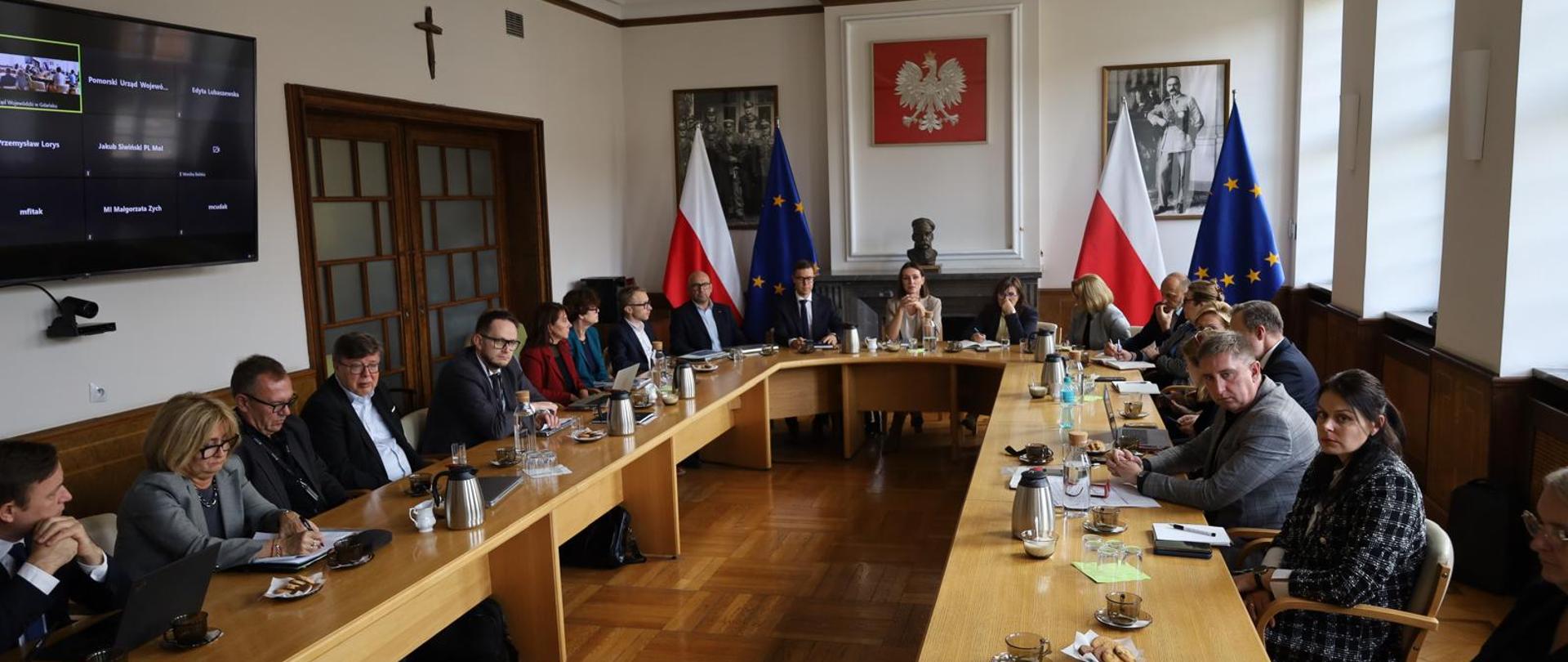 wiele osób siedzi przy okrągłym stole i dyskutuje, na ścianie wisi godło i portrety marszałka Piłsudskiego