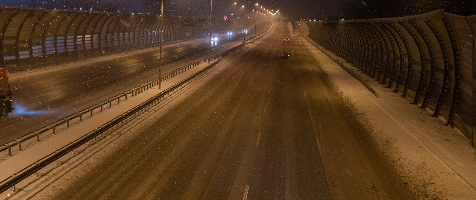 Dwupasowa droga ekspresowa w zimowej aurze. Śnieg wciąż pada. Po drodze jedzie kilka samochodów w obu kierunkach. Po bokach widoczne ekrany akustyczne.