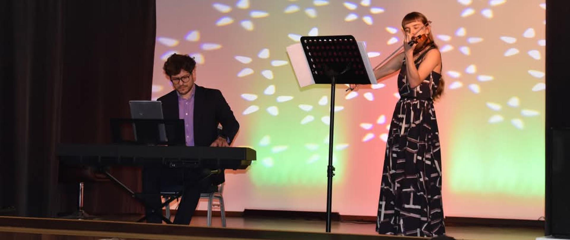 Na zdjęciu po prawej stronie dziewczyna w czarnej długiej sukience, która gra na skrzypcach. Przed nią czarny pulpit. Po lewej stronie mężczyzna brunet w okularach, fioletowej koszuli oraz czarnym garniturze, który siedzi na krześle i gra na czarnym keybordzie.W tle na ścianie rzut białych gwiazdek.