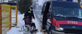 Aura zimowa, strażak OSP pomaga wysiąść z samochodu lekkiego pożarniczego z OSP Jeżów pacjentowi wyznaczonemu na szczepienie. Pacjent usiadł na wózku inwalidzkim przed wejściem do punktu szczepień.