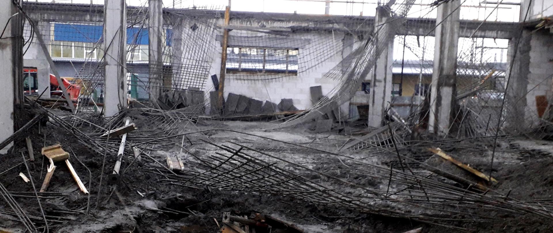 Fotografia przedstawia leżącą na podłodze połamaną i pogiętą konstrukcję stropu nad budowana halą magazynowo – produkcyjną. Widoczne są powyginane elementy metalowych kratownic wzmacniających strop oraz świeży beton porozlewany na całej powierzchni