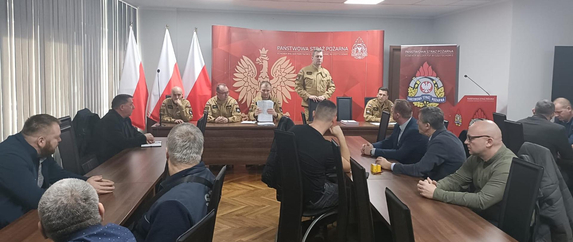 Spotkanie z przedstawicielami władz samorządowych oraz jednostek Ochotniczych Straży Pożarnych w sali konferencyjnej znajdującej się w budynku Komendy Miejskiej Państwowej Straży Pożarnej w Częstochowie. Na pierwszym planie brązowe stoły przy których na czarnych krzesłach siedzą przedstawiciele władz samorządowych oraz jednostek Ochotniczych Straży Pożarnych. Na drugim planie przy brązowym stole prezydialnym siedzą przedstawiciele Komendy Miejskiej Państwowej Straży Pożarnej w Częstochowie wraz z Komendantami. Na środku stołu prezydialnego Komendant Miejski stoi przy czarnym krześle i przemawia do osób zgromadzonych w sali. W tle czerwona ścianka reklamowa z logiem Państwowej Straży Pożarnej, białym orłem symbolizującym godło Rzeczpospolitej Polski i napisem "Państwowa Straż Pożarna, Komenda Miejska PSP w Częstochowie" po prawej baner z logo PSP a po lewej 3 flagi biało-czerwone na stojakach.