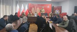 Spotkanie z przedstawicielami władz samorządowych oraz jednostek Ochotniczych Straży Pożarnych w sali konferencyjnej znajdującej się w budynku Komendy Miejskiej Państwowej Straży Pożarnej w Częstochowie. Na pierwszym planie brązowe stoły przy których na czarnych krzesłach siedzą przedstawiciele władz samorządowych oraz jednostek Ochotniczych Straży Pożarnych. Na drugim planie przy brązowym stole prezydialnym siedzą przedstawiciele Komendy Miejskiej Państwowej Straży Pożarnej w Częstochowie wraz z Komendantami. Na środku stołu prezydialnego Komendant Miejski stoi przy czarnym krześle i przemawia do osób zgromadzonych w sali. W tle czerwona ścianka reklamowa z logiem Państwowej Straży Pożarnej, białym orłem symbolizującym godło Rzeczpospolitej Polski i napisem "Państwowa Straż Pożarna, Komenda Miejska PSP w Częstochowie" po prawej baner z logo PSP a po lewej 3 flagi biało-czerwone na stojakach.