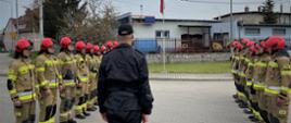 Na zdjęciu widać strażaków przed masztem na którym zawieszą flagę państwową. Uroczystość prowadzi dowódca jednostki w czarnym umundurowaniu na środku. Zdjęcie wykonane w dzień na placu jednostki. 