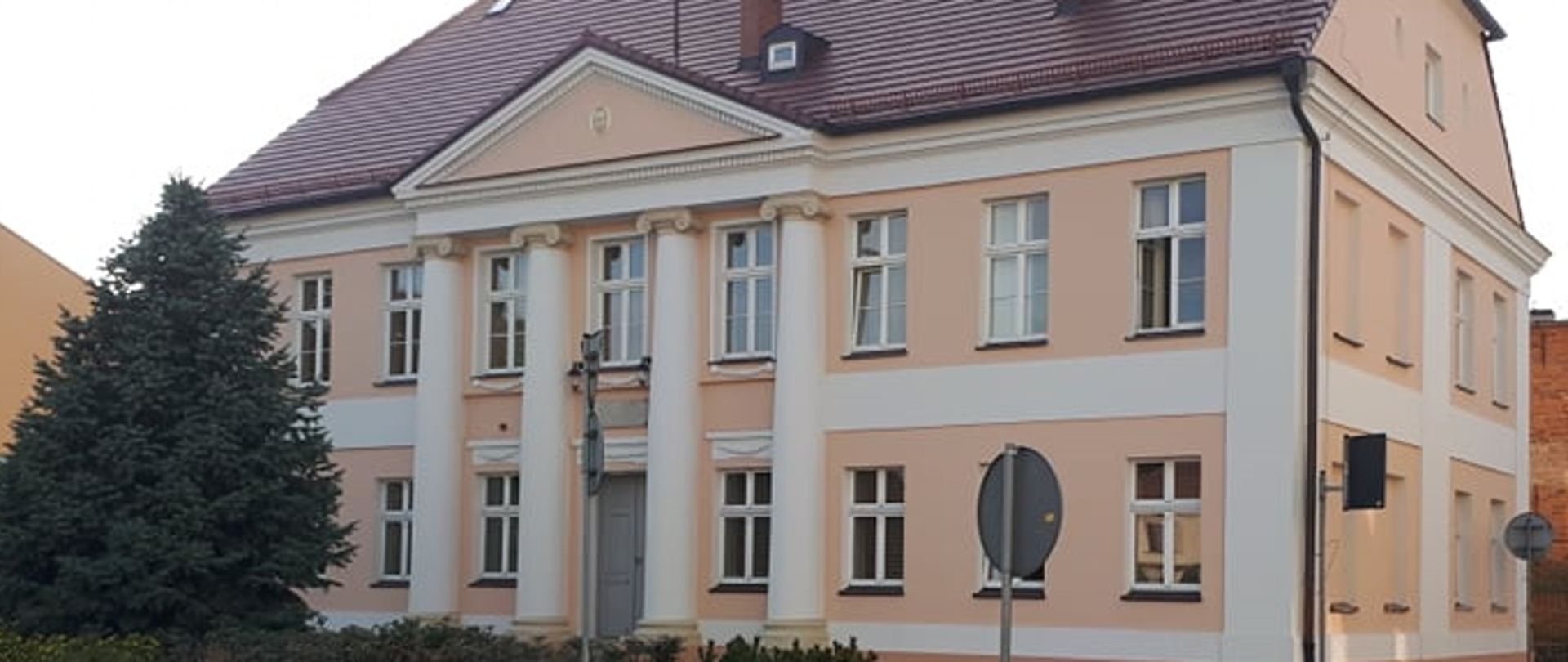 Zdjęcie przedstawia budynek PSM. Widoczne są dwie ściany budynku, front oraz ściana boczna. Na froncie znajdują się cztery kolumny rozdzielona oknami a na parterze między drugą a trzecią kolumną znajdują się drzwi główne do budynku. Przed budynkiem z lewej strony zdjęcia znajduje się wysokie drzewo iglaste a w dole na środku klomb zieleni.