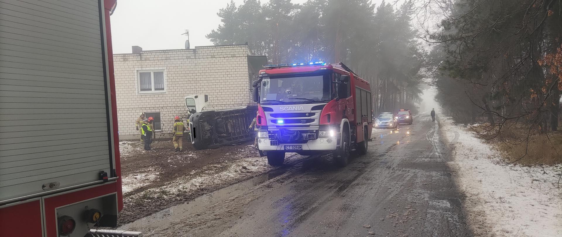Zdjęcie przedstawia pas drogowy drogi powiatowej, na której znajdują się pojazdy służb ratowniczych (dwa wozy straży pożarnej, samochód operacyjny, policja) strażaków i policjantów podczas czynności służbowych po kolizji drogowej pojazdu dostawczego przy budynku mieszkalnym z białej cegły. 