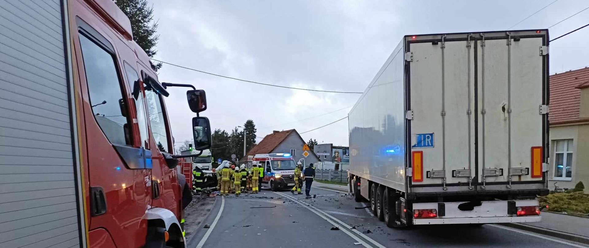 W oddali widoczne miejsce wypadku. Wokół duża ilość elementów karoserii pokrywających nawierzchnię drogi. Samochód ciężarowy uczestniczący w wypadku blokuje jeden pas drogi krajowej. Samochód osobowy blokuje drugi pas drogi krajowej. Samochód pożarniczy zaparkowany na poboczu. Trwają czynności zmierające do likwidacji skutków zdarzenia. Strażacy w ubraniach ochronnych realizują czynności na drodze.
