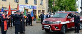 Na pierwszym planie samochód toyota hilux czerwona z napisem straż obok kapłani podczas święcenia pojazdu.